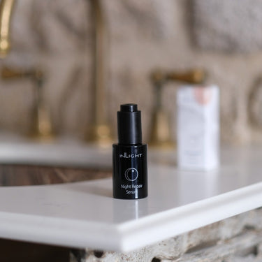 A black bottle of INLIGHT Night Repair Serum sits on a white marble countertop in a bathroom setting with a blurred stone wall and brass fixtures in the background.