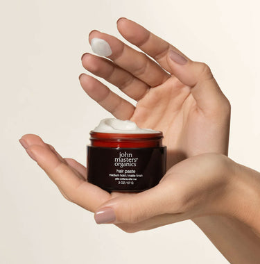 Hands holding open pot of John Masters Hair Paste showing texture