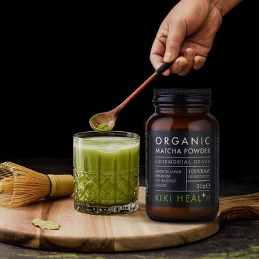 Spoon decanting matcha into a glass with the jar of Kiki Health Ceremonial Grade Organic Matcha Powder beside it
