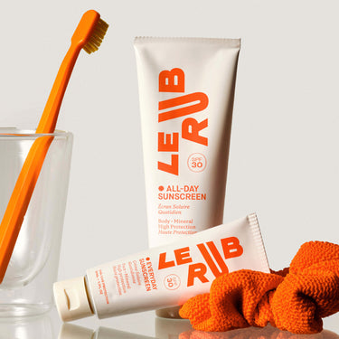 Two tubes of Le Rub The All Over SPF30 Duo on a bathroom shelf with glass holding toothbrush
